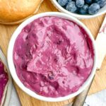 blueberry cream cheese in a white bowl on a wooden cutting board with blueberries and bagels.