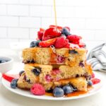 side view of a stack of sheet pan pancakes topped with berries on a white plate with syrup drizzling on top.