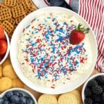 bowl of sugar cookie dip with red white and blue sprinkles on a board with cookies, fruit, and pretzels.