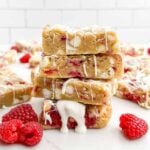 stack of white chocolate raspberry blondies drizzled with white chocolate on a white countertop with fresh raspberries.