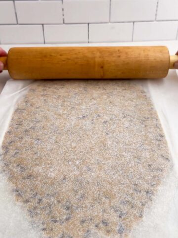 a rolling pin on a slab of chocolate chip cookie dough between two pieces of parchment paper.