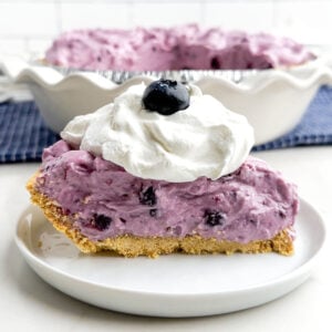 side view of a piece of blueberry cream pie on a white plate topped with whipped cream and a blueberry.