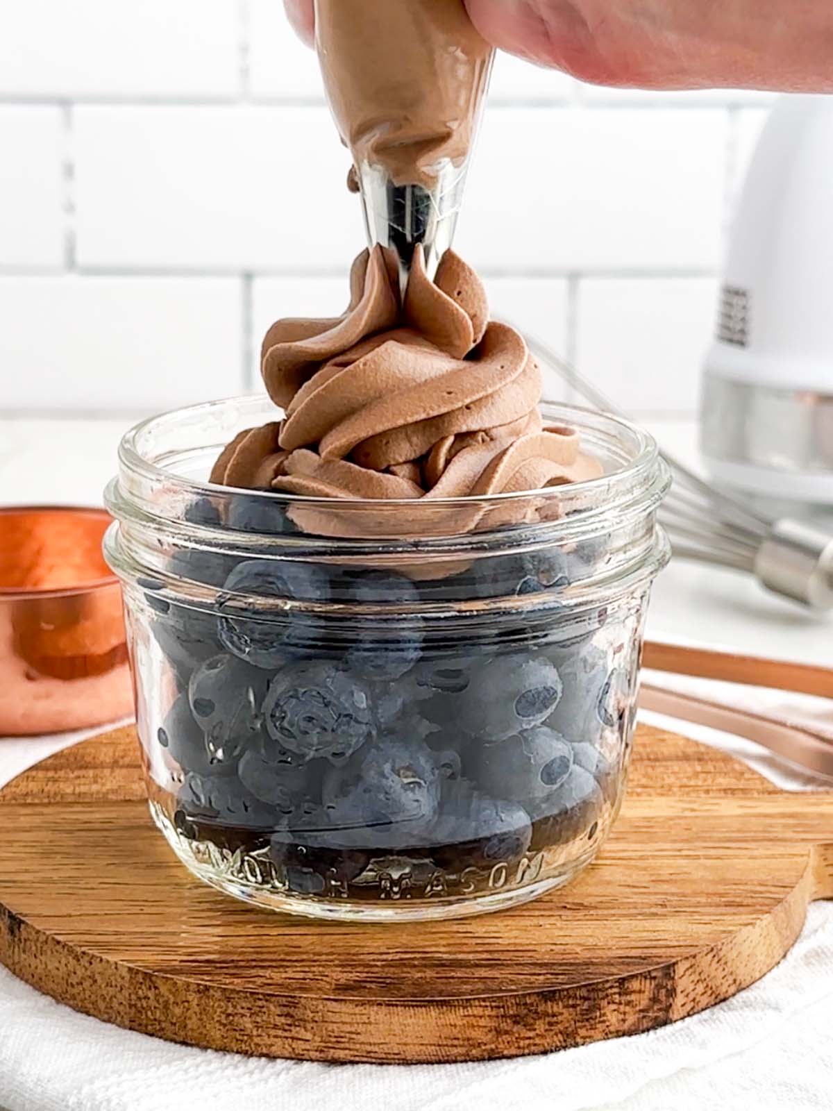 piping bag piping chocolate whipped cream on top of blueberries in a mason jar.