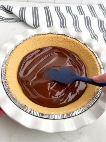 spatula spreading chocolate ganache into the bottom of a graham cracker crust.
