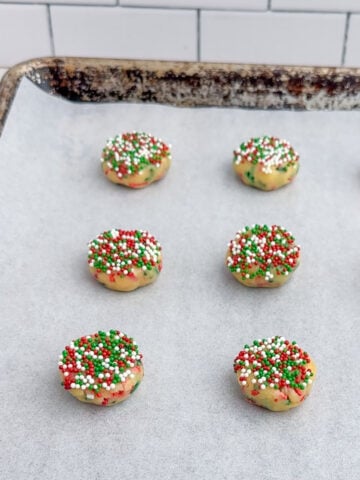 sprinkle cookie dough balls flattened and topped with sprinkles on a baking sheet.